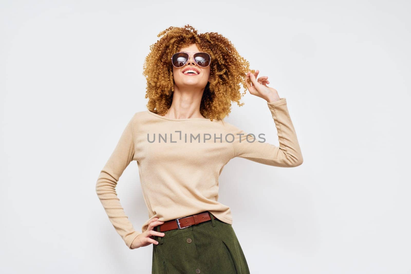 Trendy and confident young woman with curly hair wearing sunglasses, showcasing a stylish outfit in beige and green against a clean light background Perfect for fashion themes