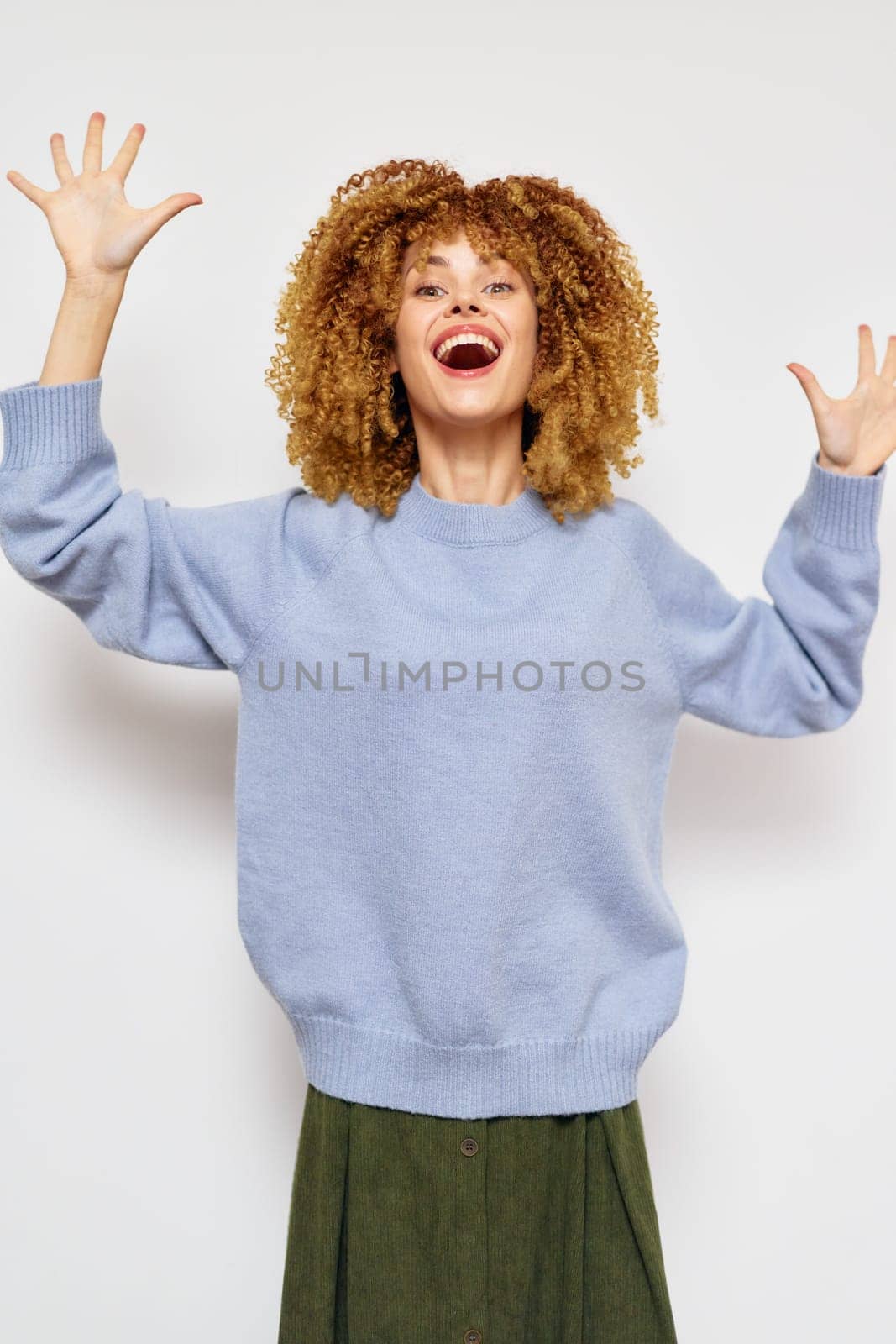 Cheerful woman with voluminous curly hair wearing a cozy blue sweater, arms raised in excitement The light background enhances her lively expression, perfect for themes of happiness and celebration