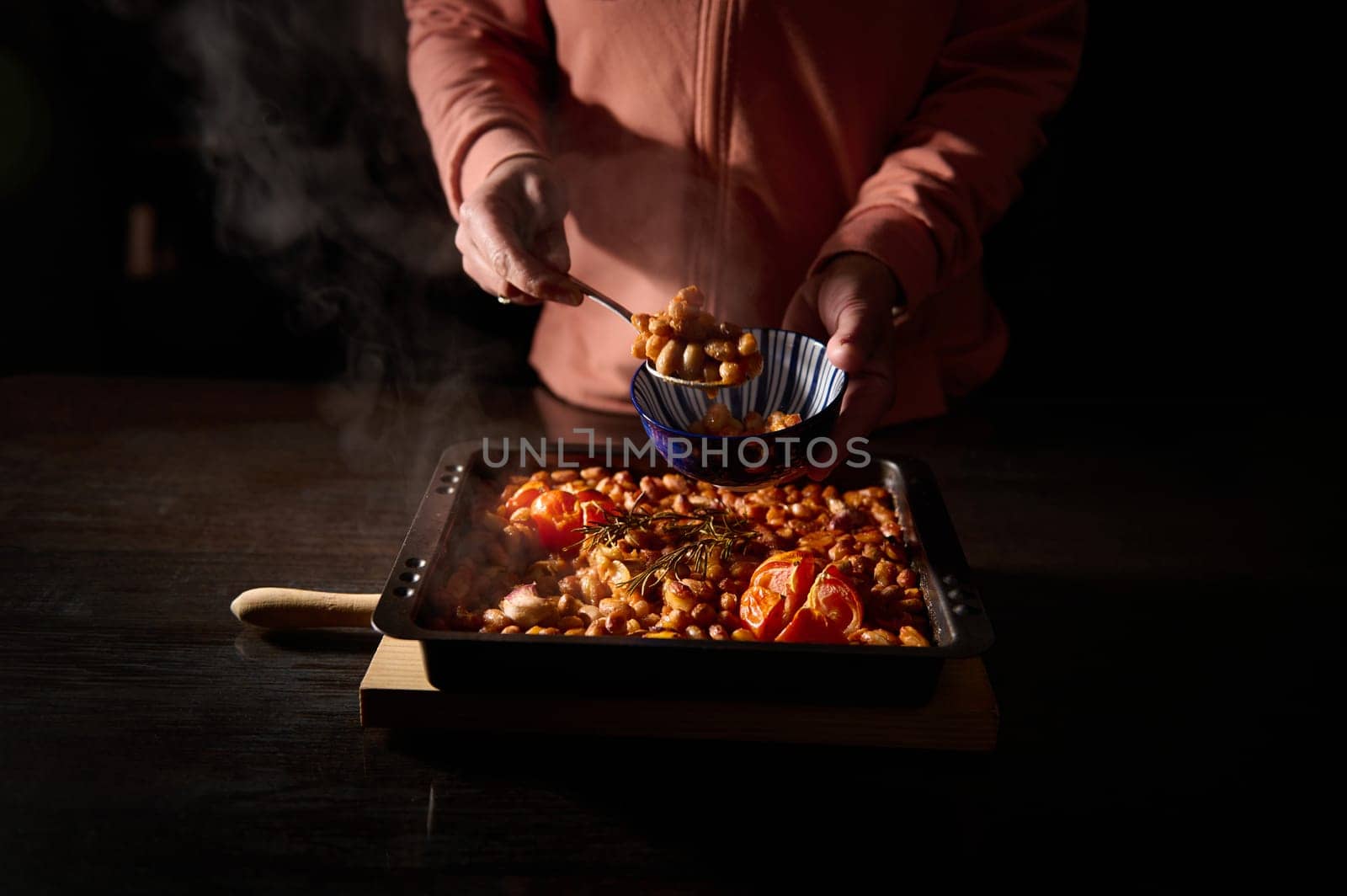 Person serving a hearty tray of baked beans and tomatoes into a bowl, set under warm, rustic lighting. A steaming tray of comforting food serves a cozy meal in an intimate setting.