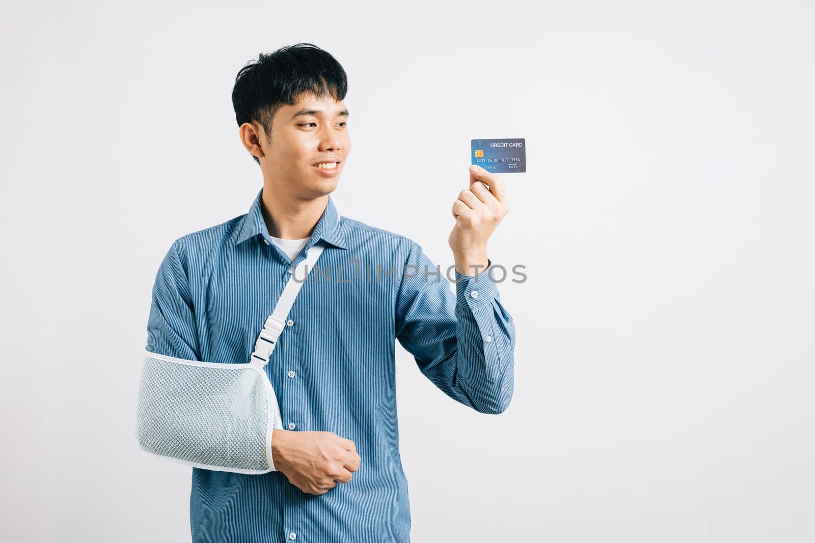 Despite a broken arm, a man wears a support splint and pays medical bills with a credit card. Happy Asian man in a sling, isolated on white, emphasizing quality health care and recovery.