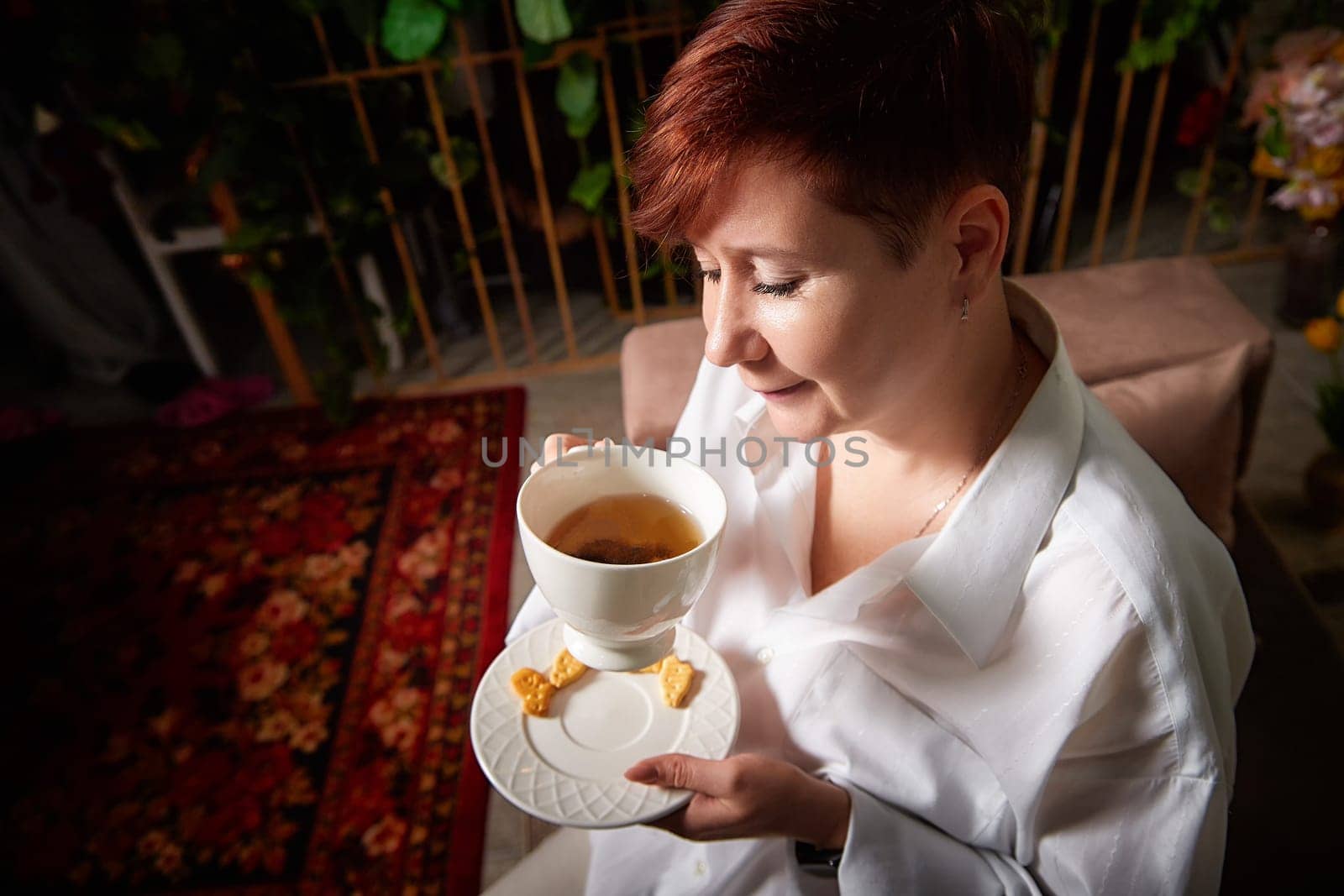 A mature woman with short hair sips tea and smiles while seated comfortably in a cozy, decorated space.