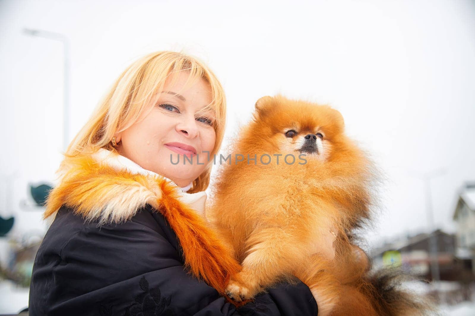 Blonde woman holds her adorable Pomeranian as they stroll through a snowy landscape, radiating warmth and joy.