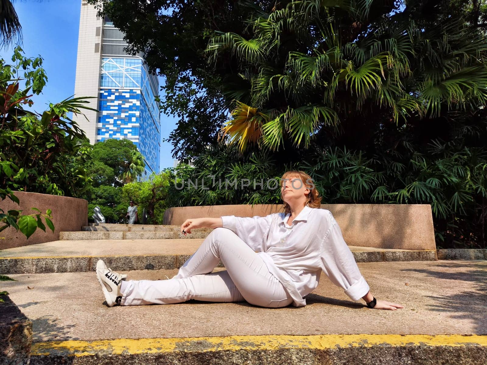Blonde woman lies back on a stone path, surrounded by greenery, while savoring a peaceful moment during her travel adventure.