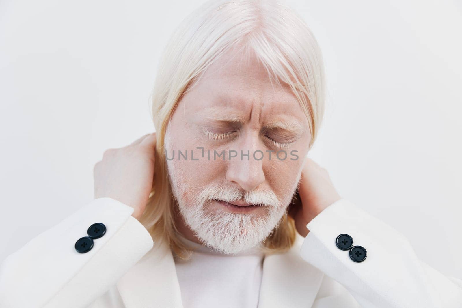 An elderly man with long white hair and a white beard dressed in a white suit shows a pained expression while holding his neck The background is a soft, neutral white, emphasizing his discomfort