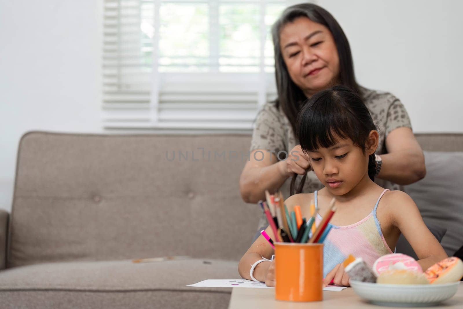 A tender moment as a grandmother styles her granddaughter's hair, reflecting love and care while the child engages in her creative drawing.