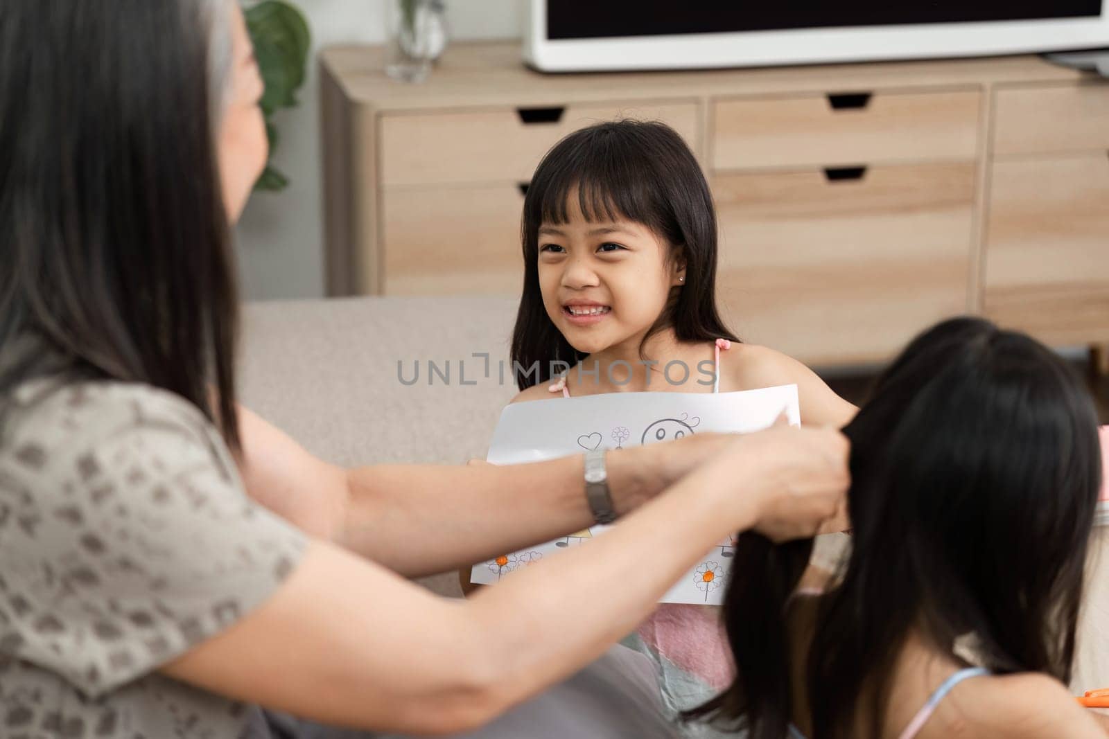A grandmother styles her granddaughter's hair, creating a nurturing moment while the child proudly displays her drawing, emphasizing family love.
