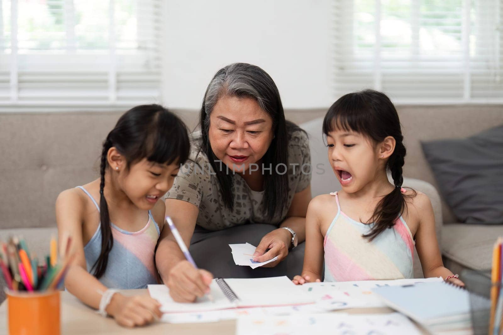 A loving grandmother assists her two granddaughters with their art projects, fostering creativity and learning in a warm family environment. The girls are excited and focused on their drawings.