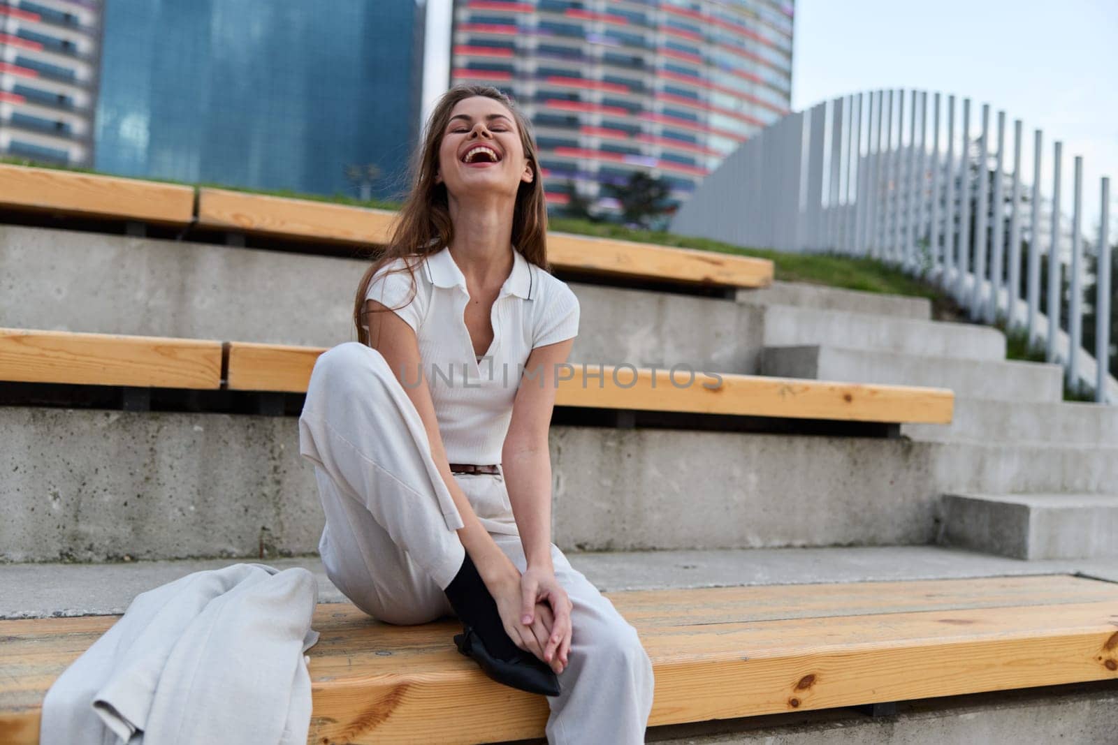 Joyful young woman in casual white clothing laughing and sitting on wooden steps in an urban outdoor setting with modern city skyline in background, expressing happiness and freedom.