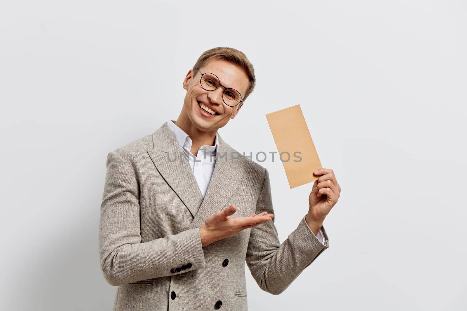 Smiling man in glasses and beige blazer holding paper card, gesturing with hand, isolated on white background. Casual business style, cheerful expression, modern communication and remote digital freelancer concept.
