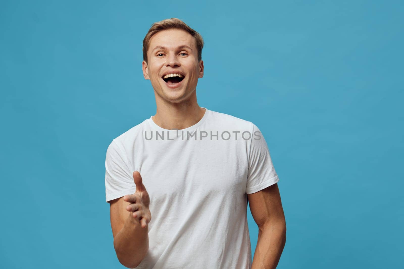 happy smiling man wearing casual white t-shirt standing against blue background showing cheerful joyful facial expression with open mouth and laughter in studio portrait. Concept: positive emotion and happiness