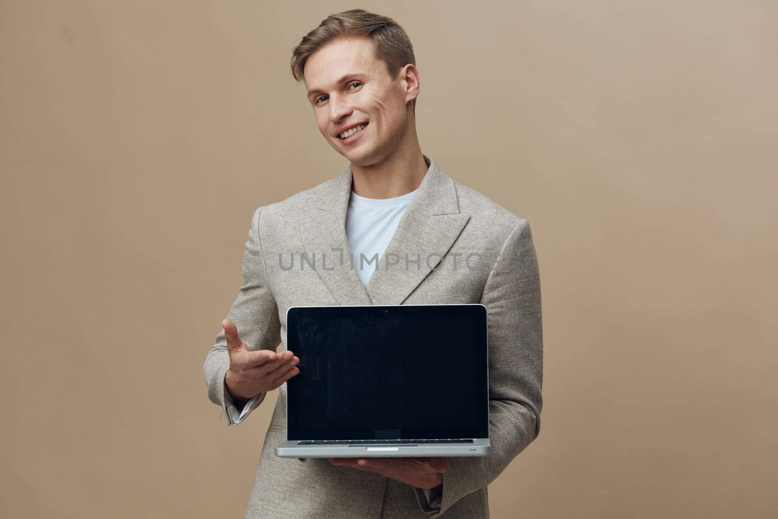 smiling young man holding laptop with blank screen wearing beige blazer standing isolated on beige background. The image represents digital nomad lifestyle, freelance work, and remote job concept involving modern technology and mobility. Concept: freelance and digital nomad