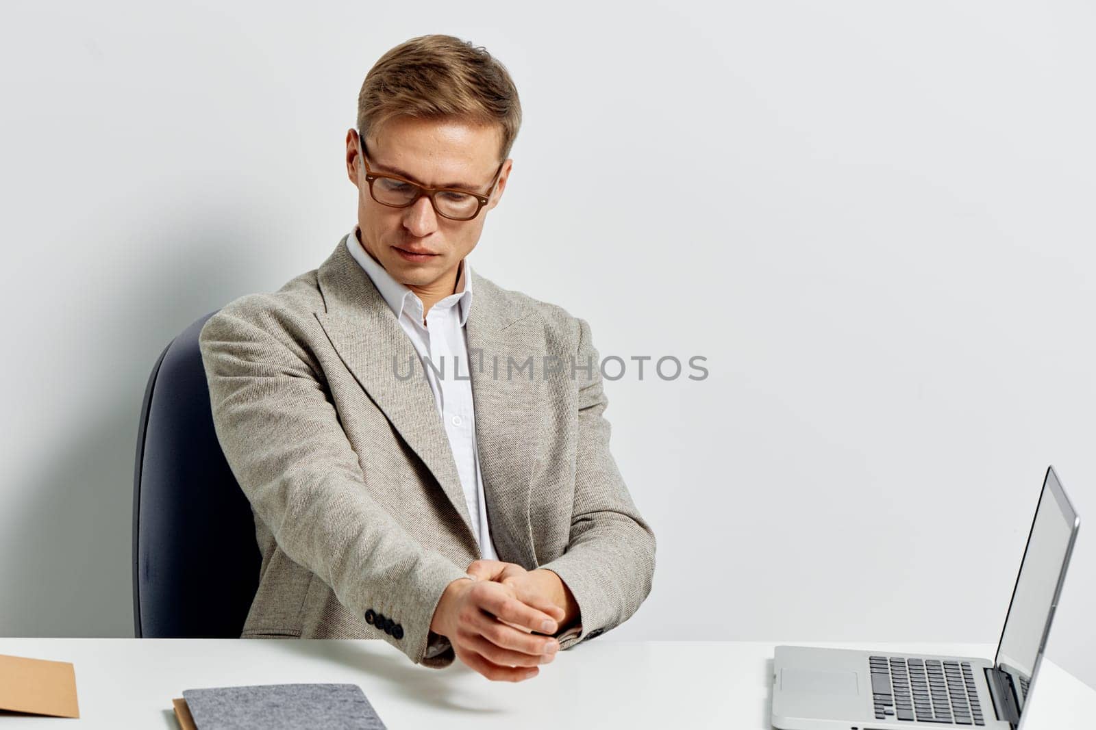 young man stretching hands while sitting at white desk with open laptop and notebook wearing casual blazer and glasses focused on work digital nomad lifestyle and remote work in metaverse concept