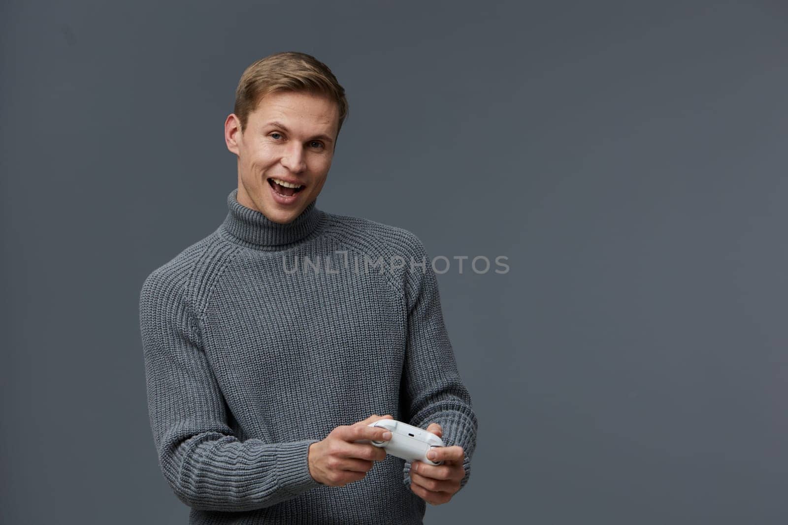 cheerful young man holding game controller in hands wearing gray sweater playing video game with excitement on face isolated on gray background indoor casual clothing digital entertainment concept