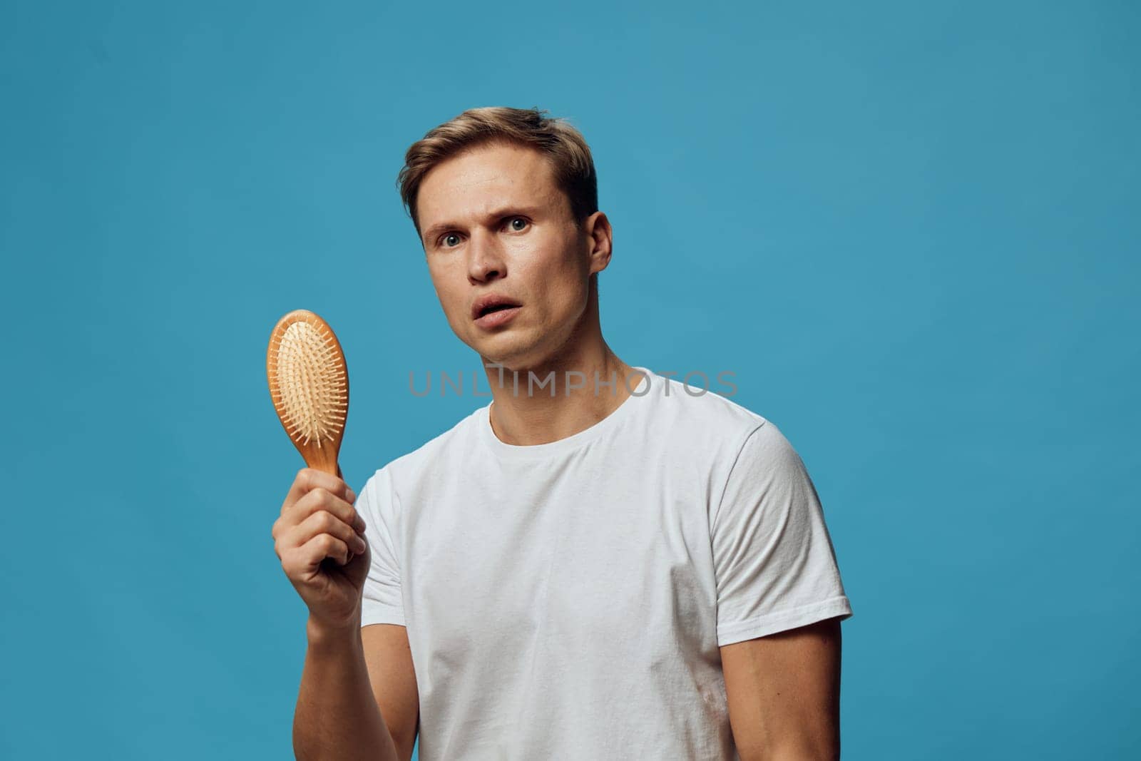 young caucasian man wear plain white t-shirt hold wooden hairbrush look camera with confused skeptical expression on blue background mental health and gadgets concept focus on emotions and self-care