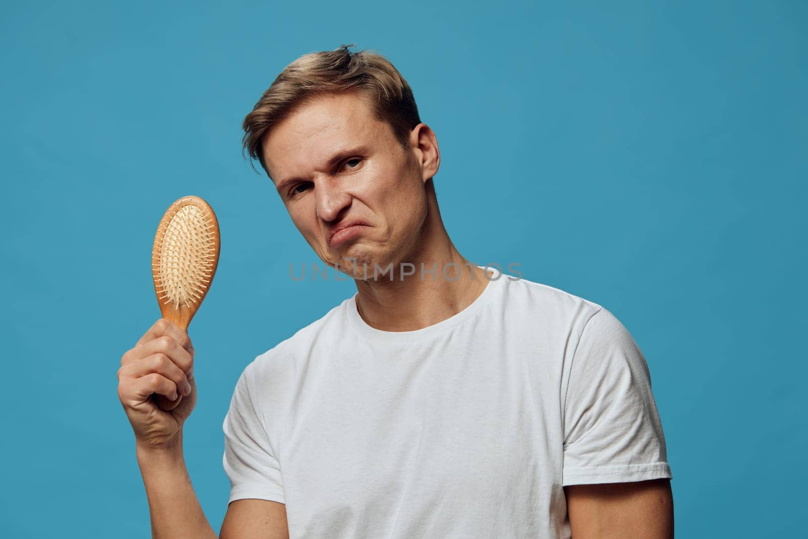 unhappy man holding hairbrush wearing white t-shirt looking displeased against blue background, facial expression showing dissatisfaction and frustration, male grooming and personal care theme, concept: mental health and self-image awareness
