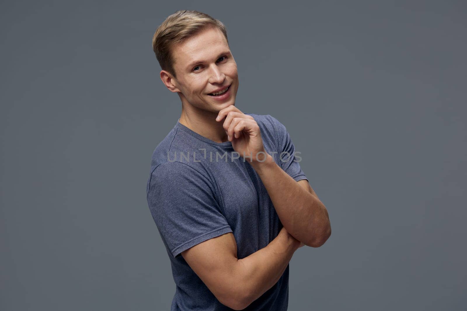 smiling young caucasian man in blue t-shirt pose thinking with hand near chin look at camera isolated on grey background. Relaxed confident digital nomad work concept with casual clothing and short hairstyle. Concept: digital nomad lifestyle