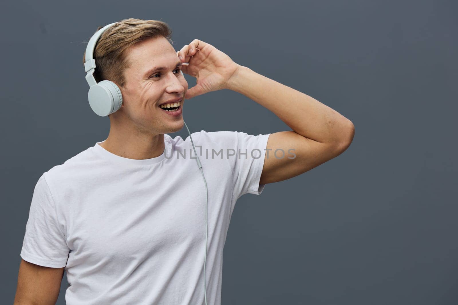 Smiling young man wearing white headphones and casual white t-shirt looking sideways while listening to music or participating in video calls. Modern gray background enhances focus on cheerful expression and relaxed pose. Concept: digital nomad lifestyle