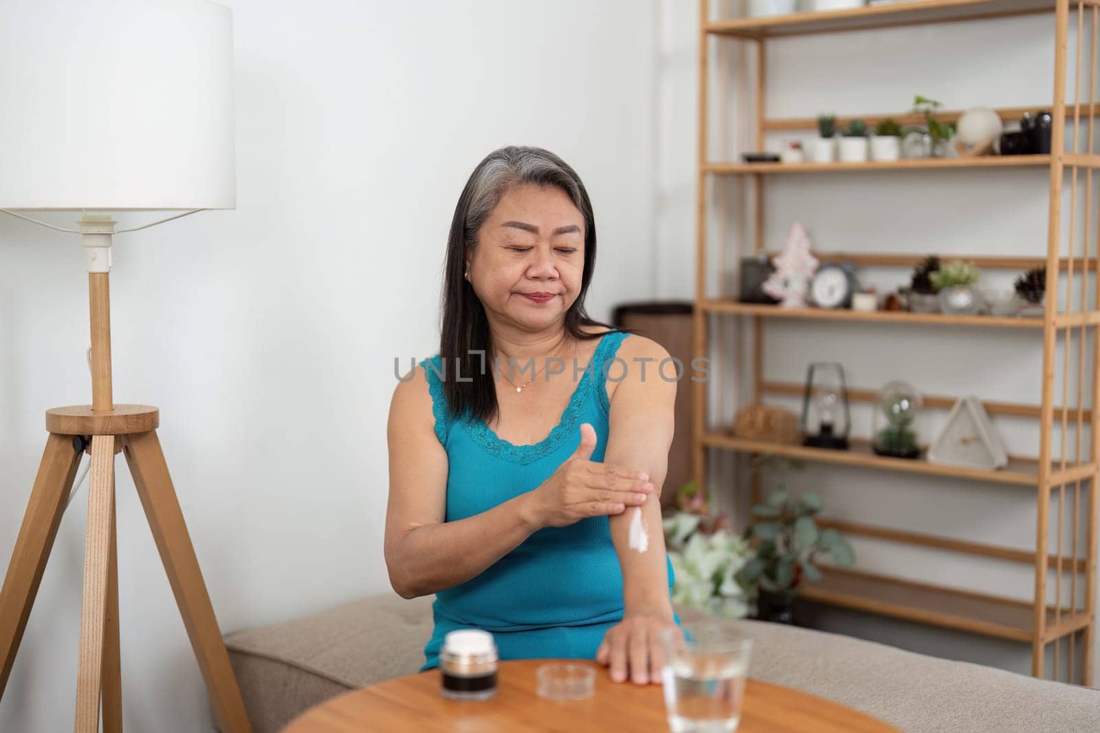 An elderly woman enjoys a moment of self-care, applying lotion to her arm while seated comfortably at home. This image highlights the importance of beauty and wellness routines at any age.