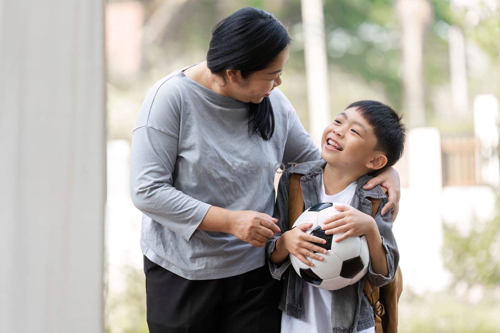 A mother smiles at her son as he holds a soccer ball, showcasing their bond and excitement for outdoor activities together.