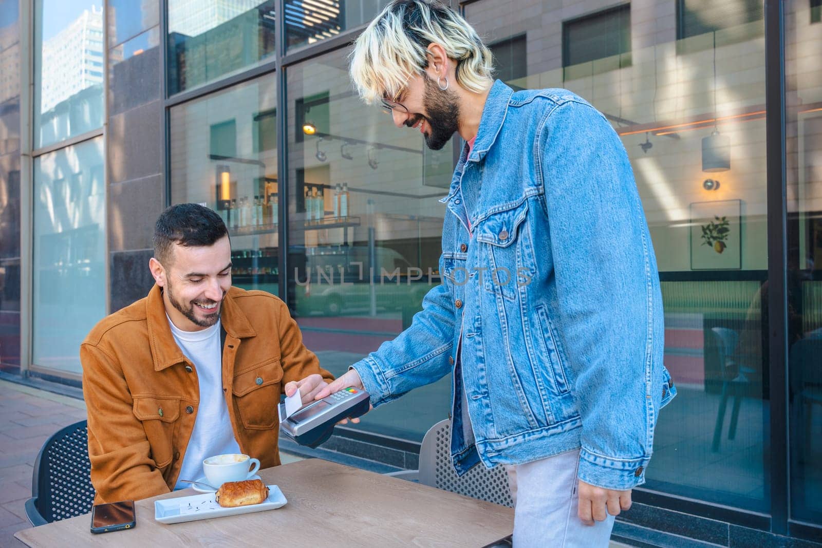 Caucasian Man pays with a contactless card at an outdoor cafe while his friend watches and smiles. Modern street cafe scene with relaxed atmosphere and casual fashion in an urban environment.