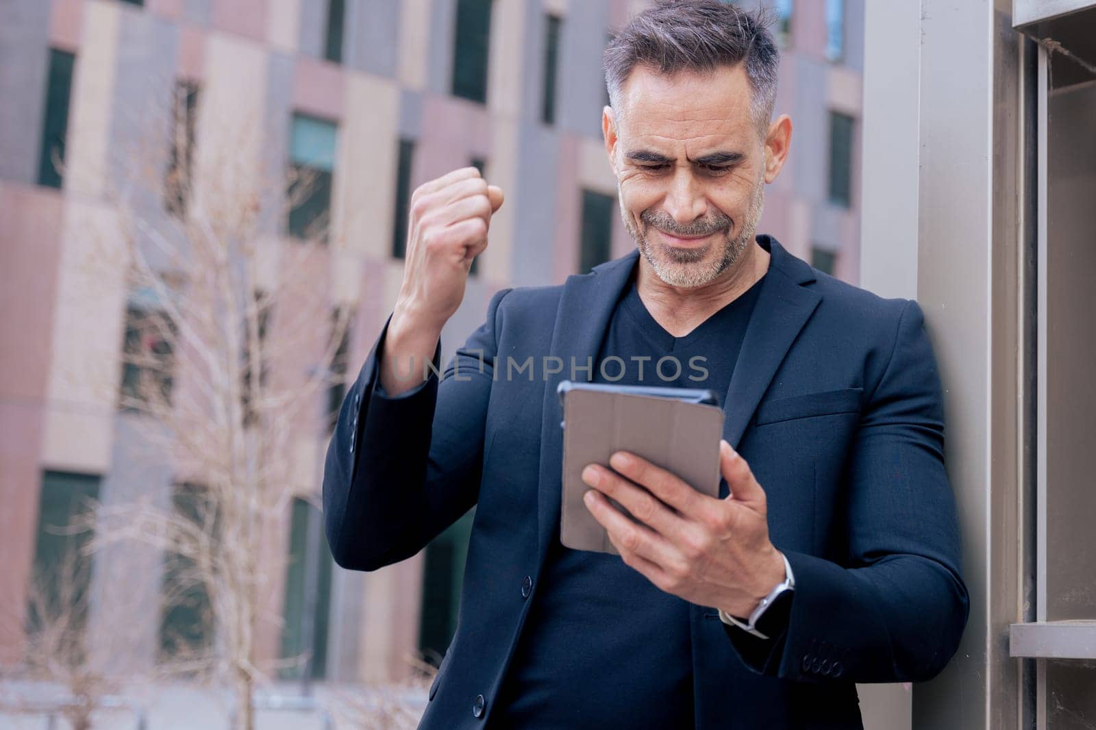 Confident businessman celebrates winning an online bet, holding a tablet and raising his fist. He smiles outdoors in a city setting. Concept of success, digital lifestyle, and modern finance.