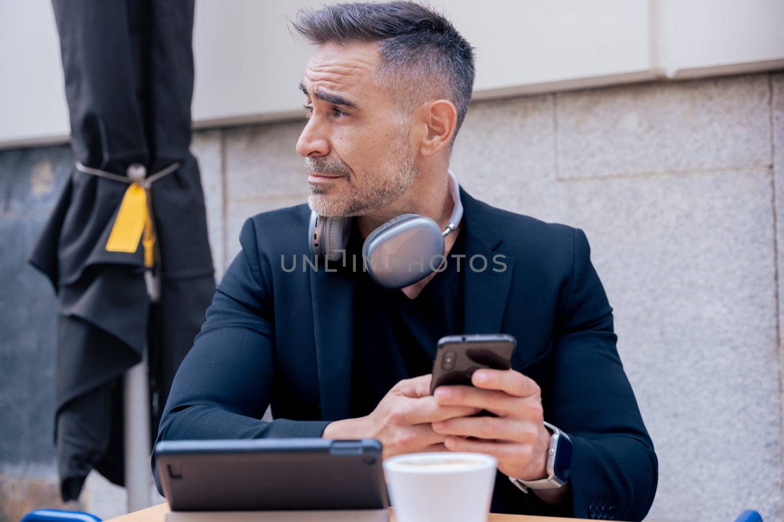Mature businessman with headphones around neck sits at a cafe table holding a smartphone. Tablet and coffee on the table. He looks aside with a thoughtful expression in a modern urban setting.