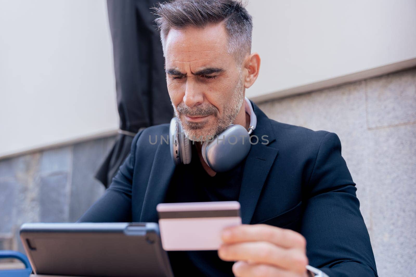 Confident caucasian man with headphones smiling while using tablet and holding a credit card outdoors. Concept of online shopping, digital payment, modern lifestyle, and financial technology.