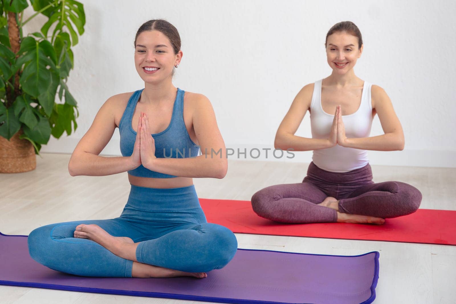 Caucasian women practicing yoga in seated lotus pose with palms together, wearing sportswear on mats in a bright indoor space. Calm expression and focus reflect mindfulness and healthy lifestyle