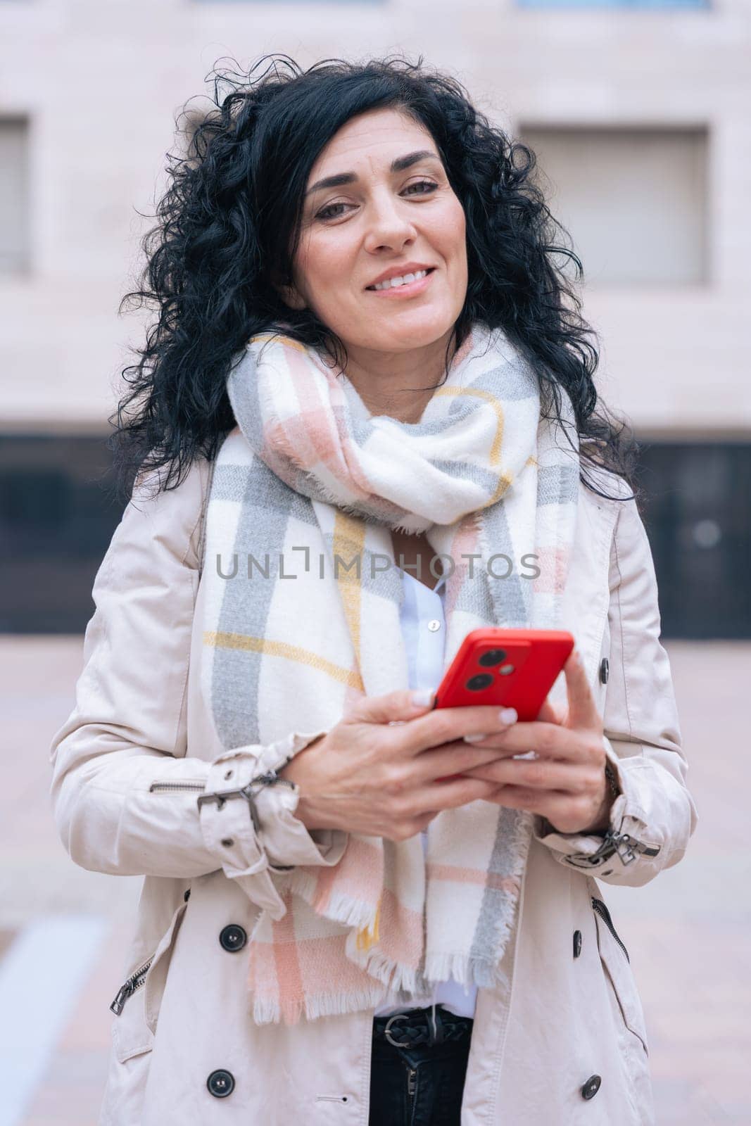 Happy caucasian woman standing outdoors in urban setting,wearing coat and scarf,using her red smartphone.She is smiling and relaxed,enjoying a casual moment on a cold day in the city looking at camera