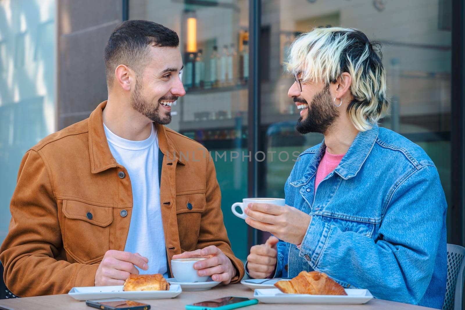Two friends share a joyful moment at an outdoor cafe, smiling while drinking coffee and eating pastries. Casual conversation and relaxed vibe in an urban setting with natural light and glass walls.