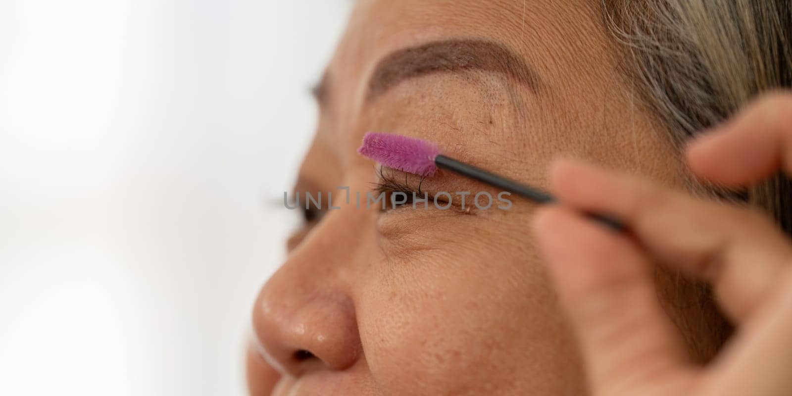 An elderly woman carefully brushes her eyebrows, highlighting her attention to detail in her beauty regimen and self-care.