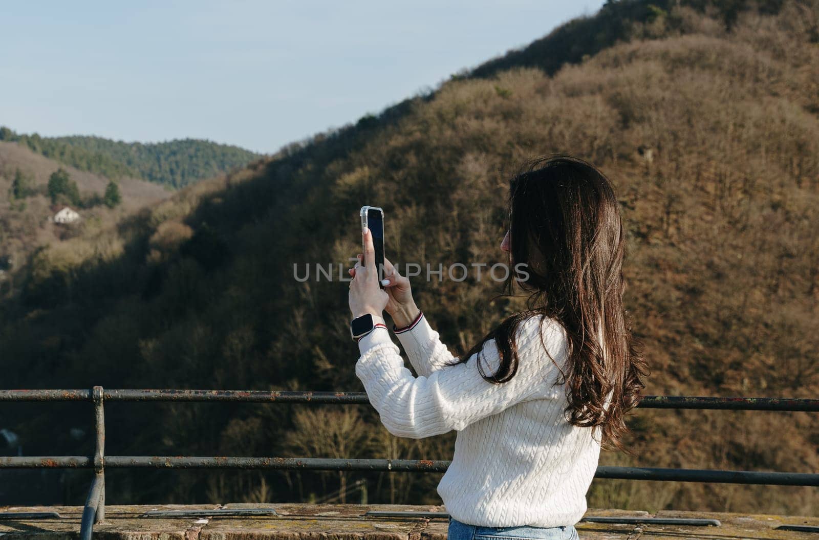 Portrait from the waist up sideways of a young beautiful brunette girl who is filming a mountain landscape with a mobile phone standing on a viewing platform in an ancient castle, a spring sunny day, a side view close-up. Concept using technology, time of travel and tourism.