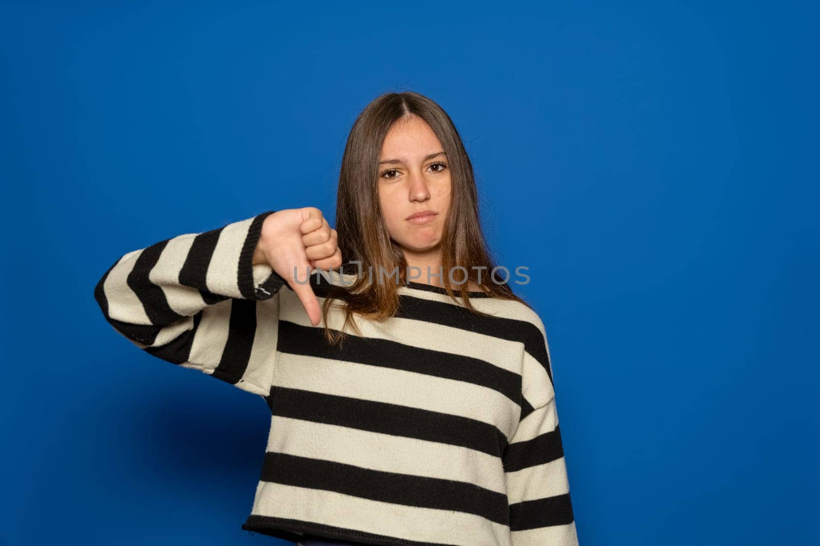 Young brunette Hispanic teenager wearing a striped sweater looking unhappy and angry, showing rejection and negativity with a thumbs-down gesture, isolated over blue background. Bad expression