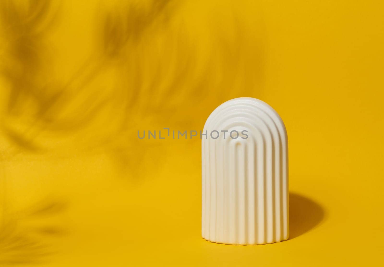 A small, white ceramic decorative arch with vertical ribbed texture stands on a bright yellow surface with blurred leaf shadows.