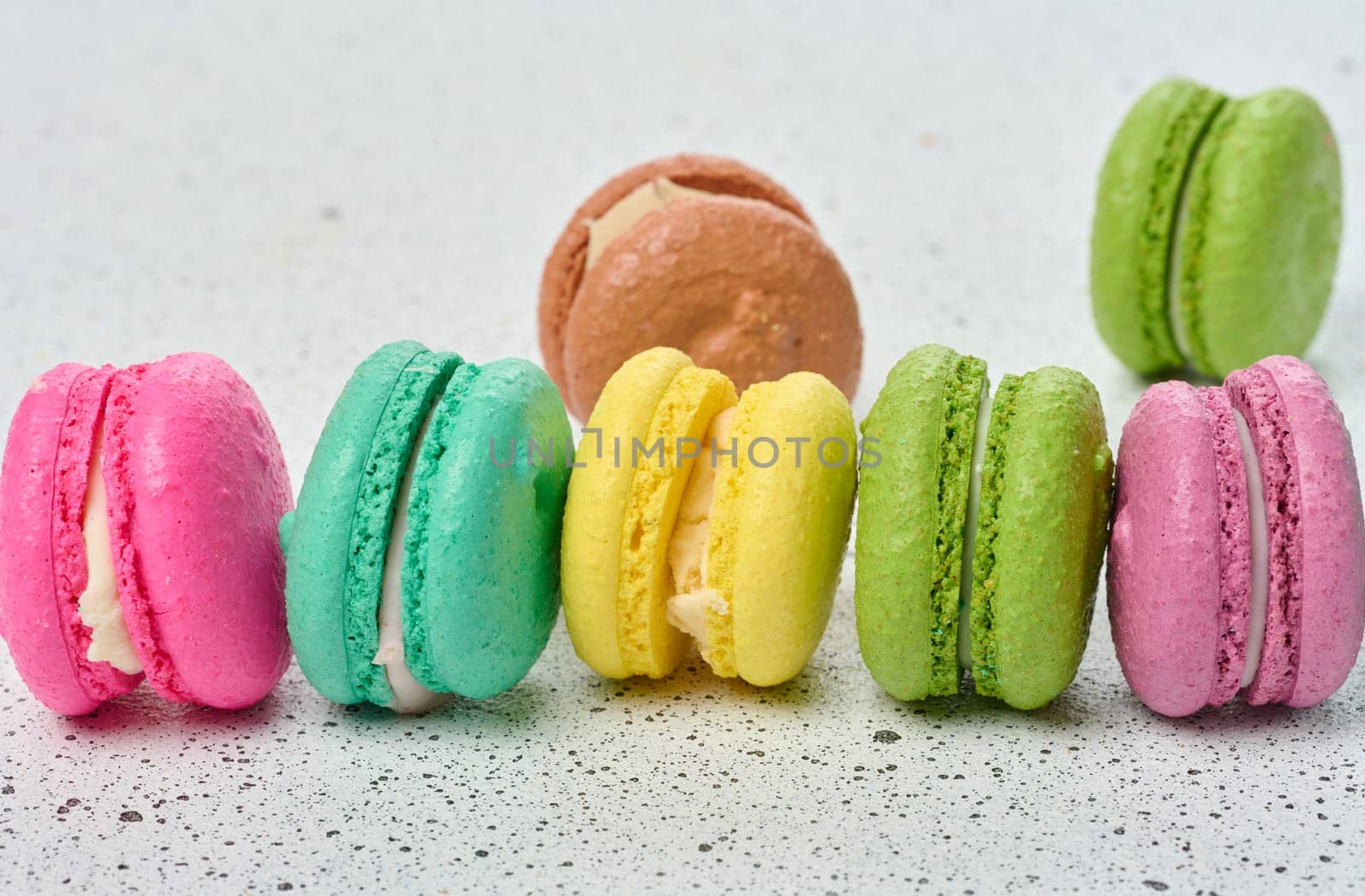 An assortment of colorful macarons on a white surface with dark speckles. The macarons have a smooth, delicate shell and a creamy filling