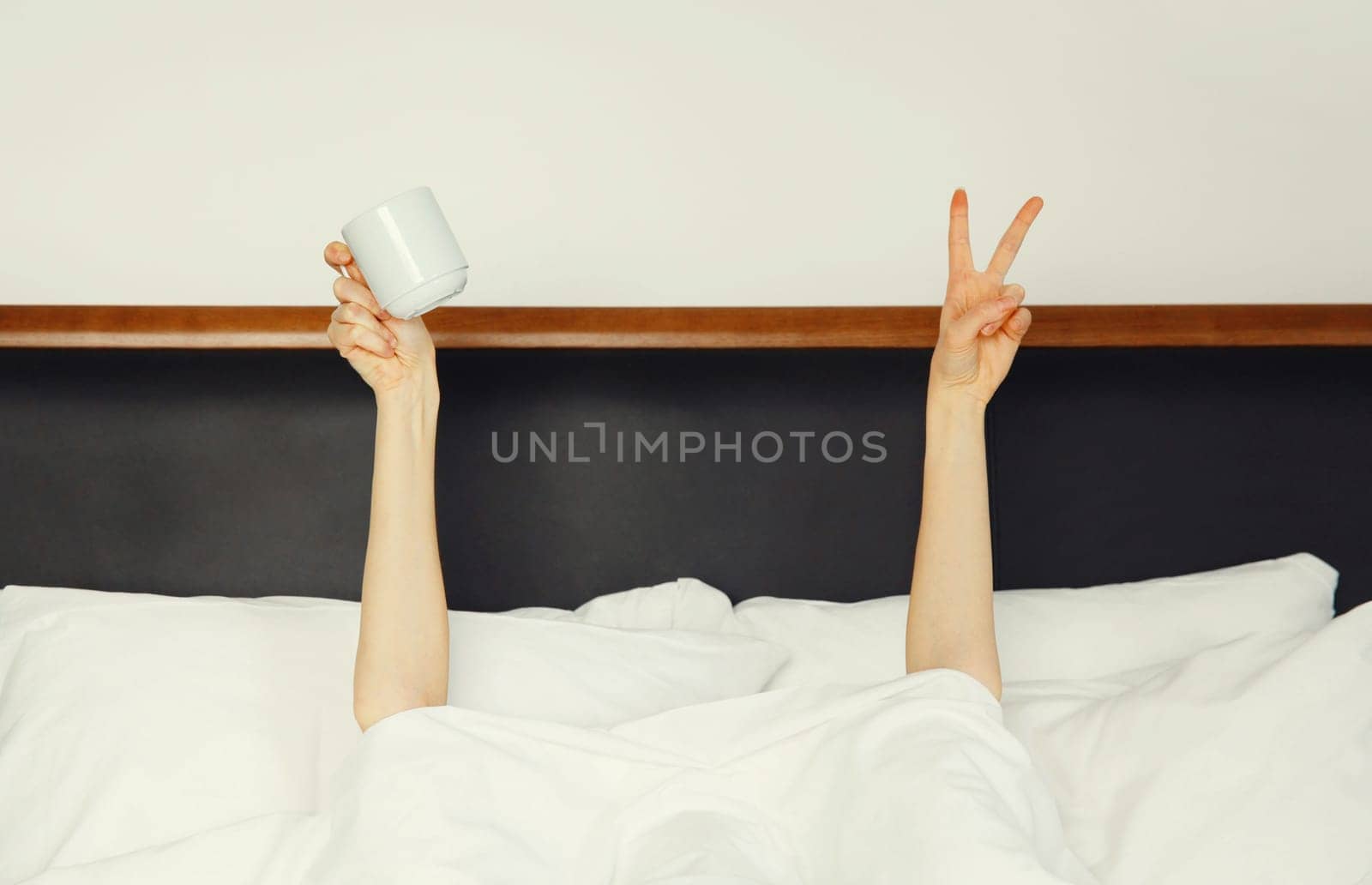 Cheerful lazy woman waking up after sleeping in the morning while lying in white soft comfortable bed showing empty cup coffee stretching her hands up from under the blanket in bedroom at home