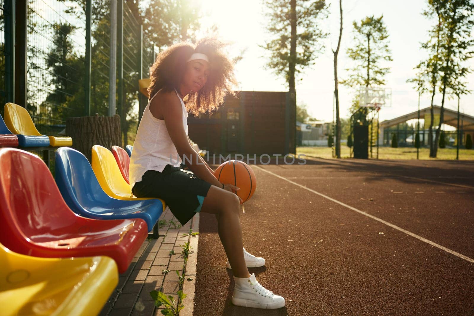 Attractive confident young sportswoman with basketball ball in hands sitting taking rest during training class at outdoor court