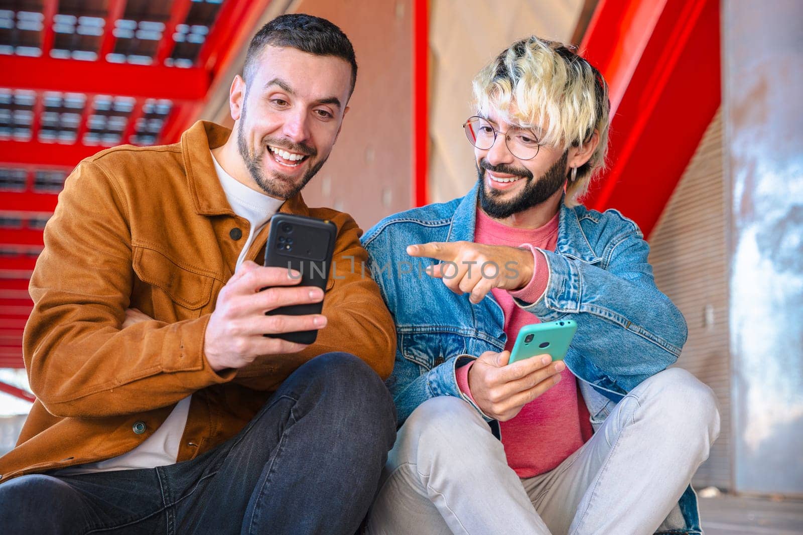Two friends sit together outdoors, laughing and pointing at a smartphone screen. Casual outfits and red architectural background suggest a modern, social and relaxed moment in the city.
