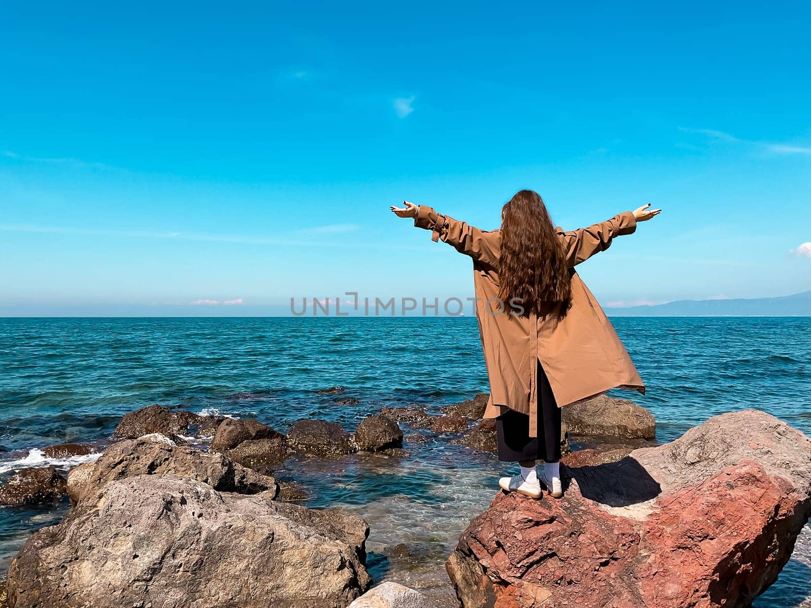 Young caucasian female enjoying ocean view on rocky shore under clear blue sky.