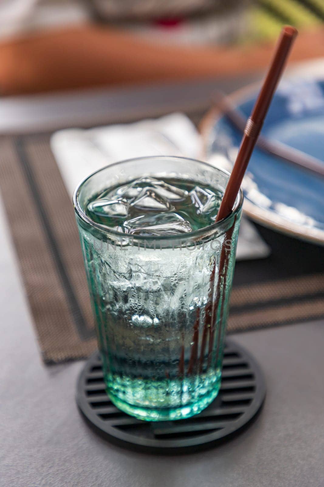 Water and ice in a clear glass with a brown plastic straw and a black coaster. In a restaurant, an empty plate in a blurred background. Selective focus.