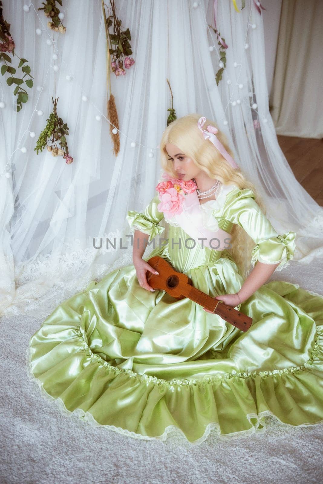 young blonde with a small guitar in her hands, and in a medieval dress, in the Baroque style, sits on a white background made of fabric. dress of soft green color with a pink ribbon and pink flowers.