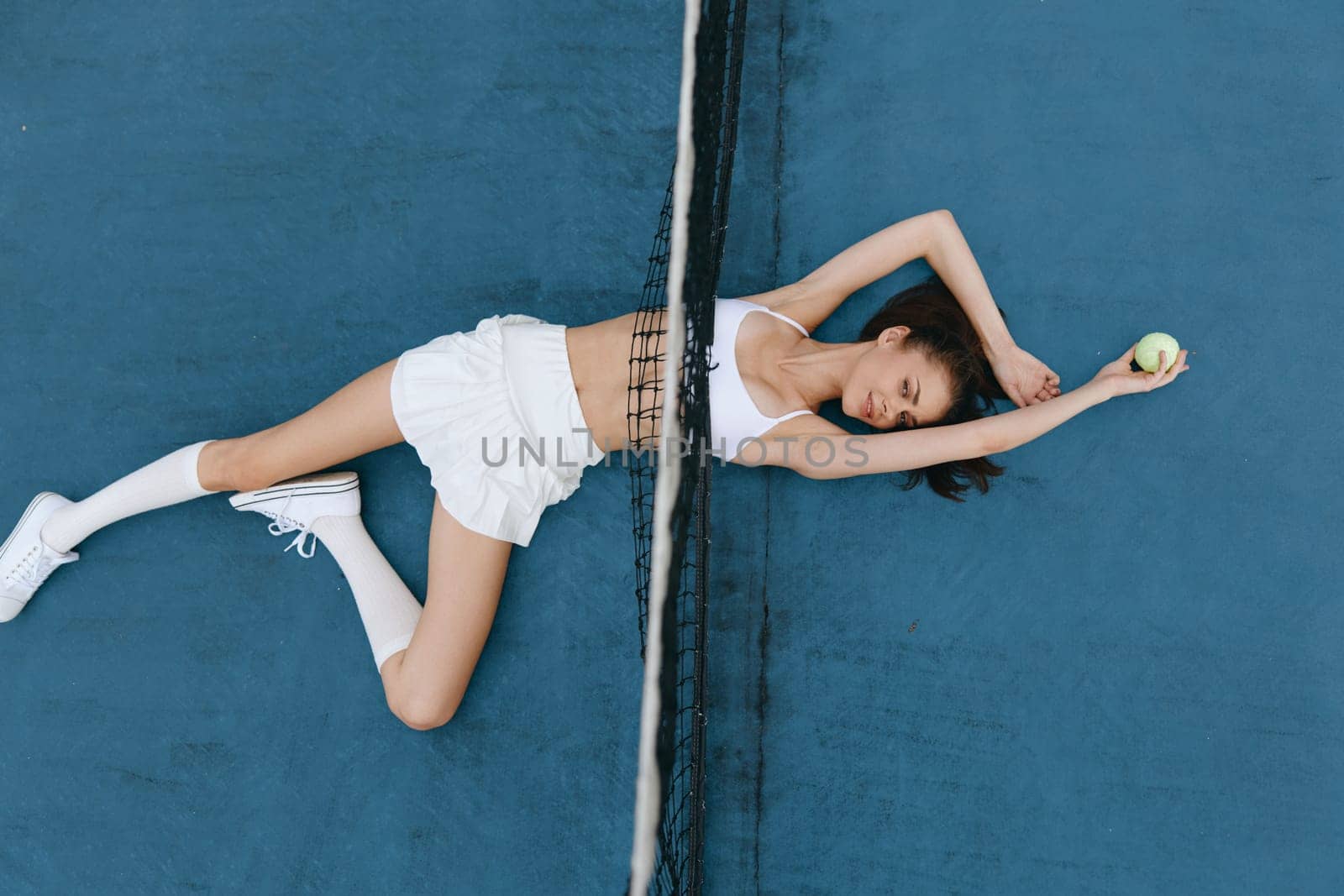 Confident young woman in white sportswear lying on a vibrant blue tennis court, embodying relaxation and athleticism while holding a tennis ball, perfect for sports or wellness themes