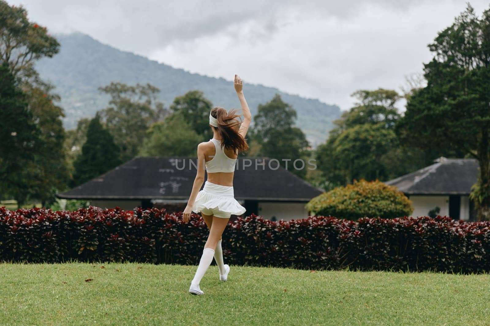 A cheerful young woman in a white outfit twirls in a lush green park, surrounded by nature and mountains, radiating joy and freedom