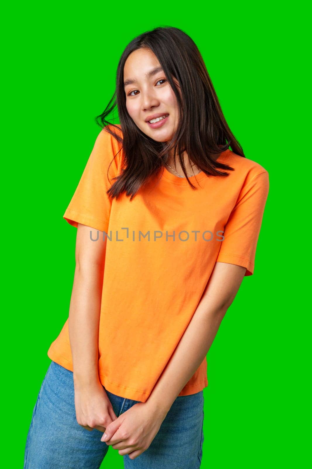 A young woman stands with a cheerful expression, wearing an orange shirt and denim jeans. She is smiling and appears to be enjoying the moment, with a lively green backdrop.
