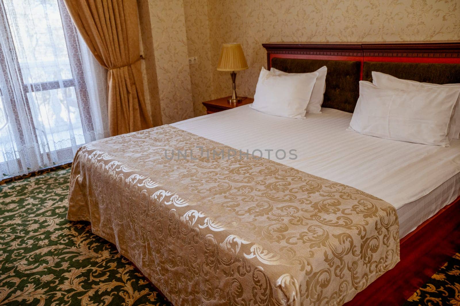 Hotel room with golden yellow blanket on dark red bed and green carpet on the floor. Room in golden beige tones with white bed linen. on the bedside table table lamp