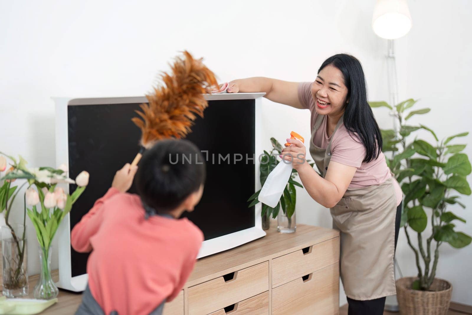 A mother and her son share a joyful moment while cleaning a TV stand, demonstrating teamwork and teaching valuable life skills in a cozy home environment.
