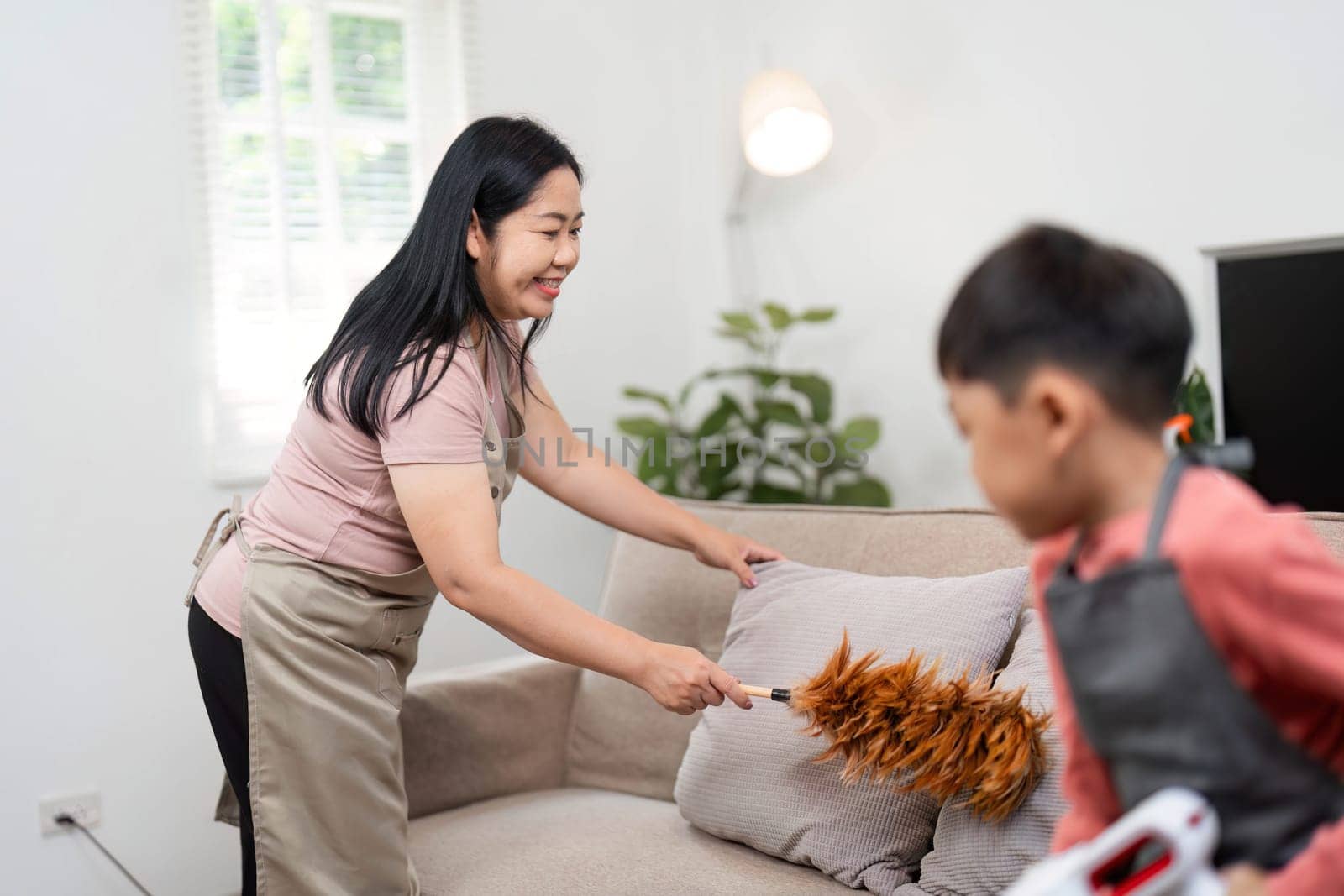 A cheerful mother engages her son in cleaning the living room, using a duster while he operates a vacuum, showcasing teamwork and fun.
