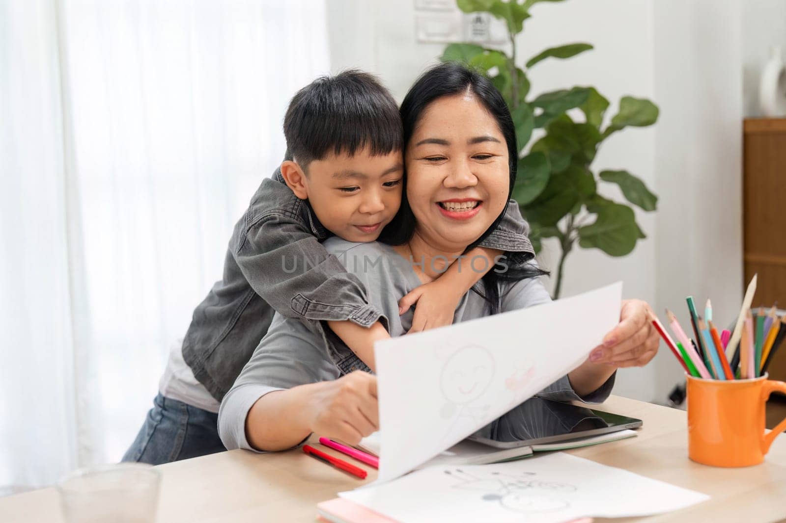 A mother and her son share a loving moment as he hugs her from behind, both delighted by his creative drawing, showcasing their close relationship.