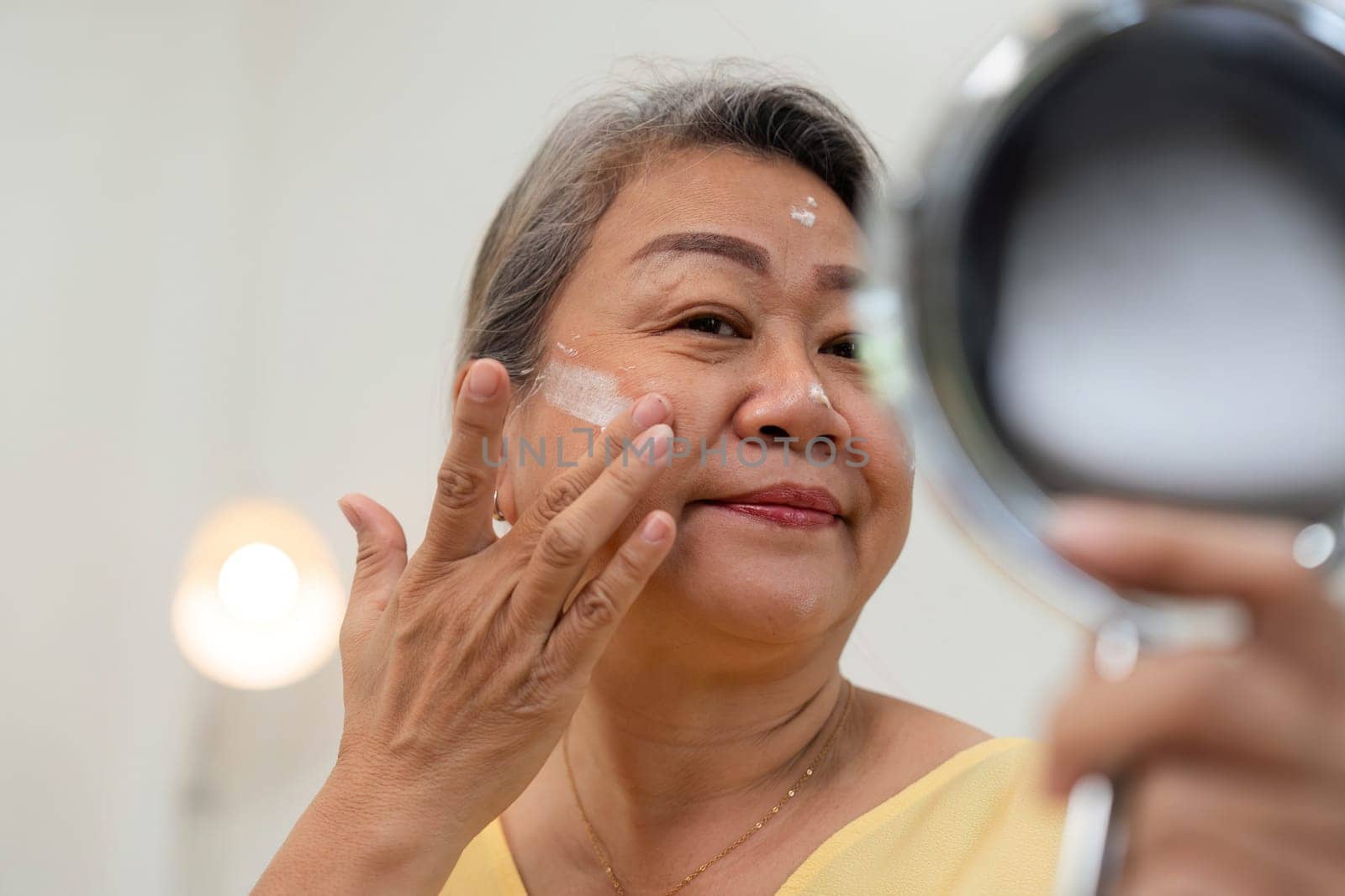A senior woman smiles as she applies skincare, reflecting joy and confidence in her beauty routine. This image embodies age diversity and self-care.
