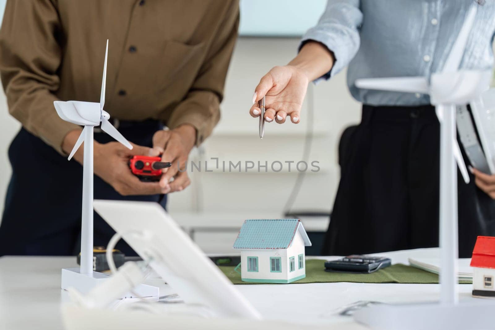 In a bright office, two professionals discuss their sustainable design project, highlighting renewable energy solutions with models of homes and wind turbines.